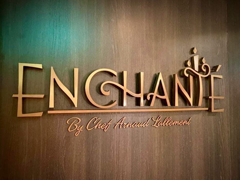 Gold Enchanté signage reading 'Enchanté by Chef Arnaud Lallement' mounted on a wood-paneled wall